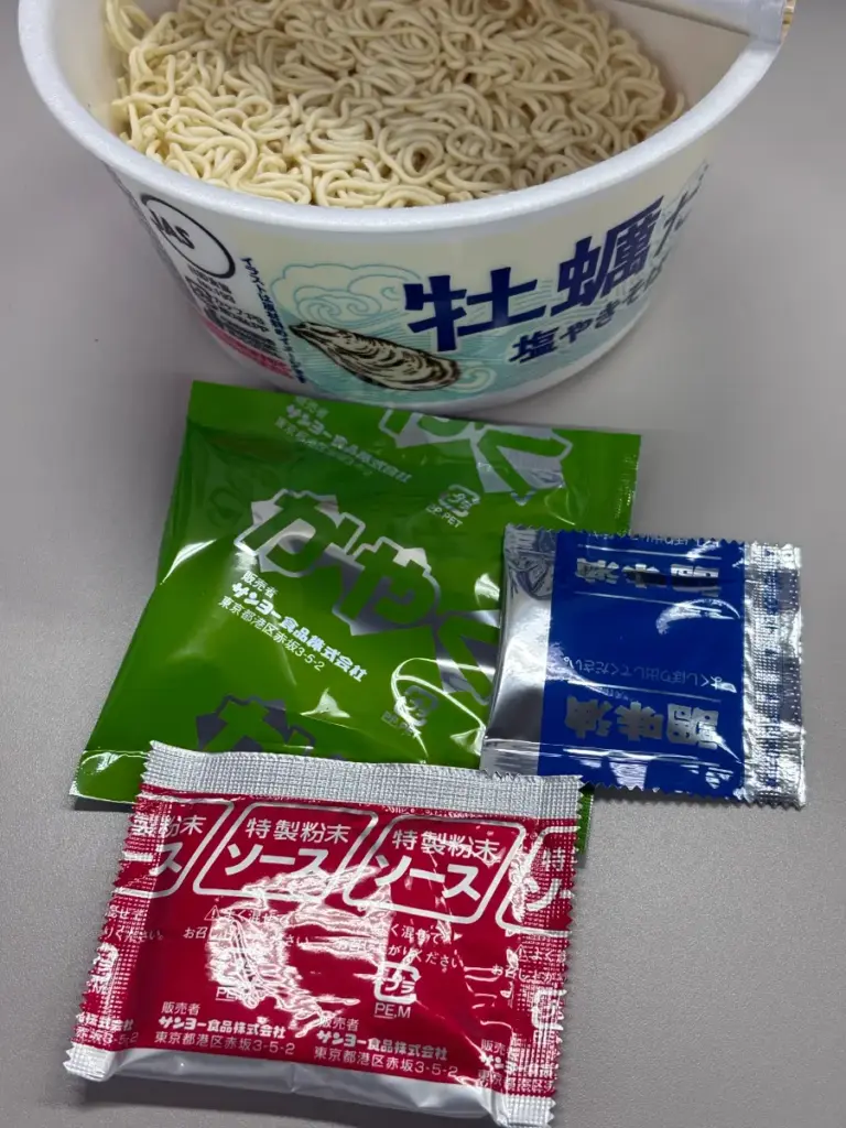 KALDI 牡蠣だし塩やきそばの中身と小袋セット。乾麺が入ったカップと、かやく(緑)、粉末ソース(赤)、後入れ調味油(青)の3種類の袋が並ぶ。調理前の証拠写真としてレビュー・評価記事に使用。