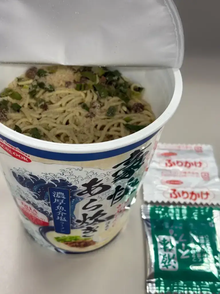 エースコック「まぐろのかぶとだし仕立て 濃厚魚介塩ラーメン」調理前。具材と粉末スープが麺の上に見え、ふりかけ小袋が添えられている。
